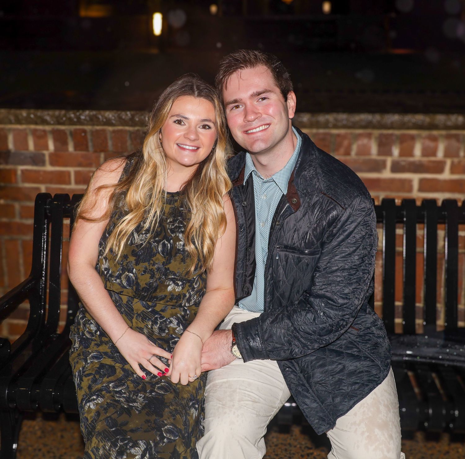 A couple in evening attire sitting together on a bench against a brick wall backdrop.