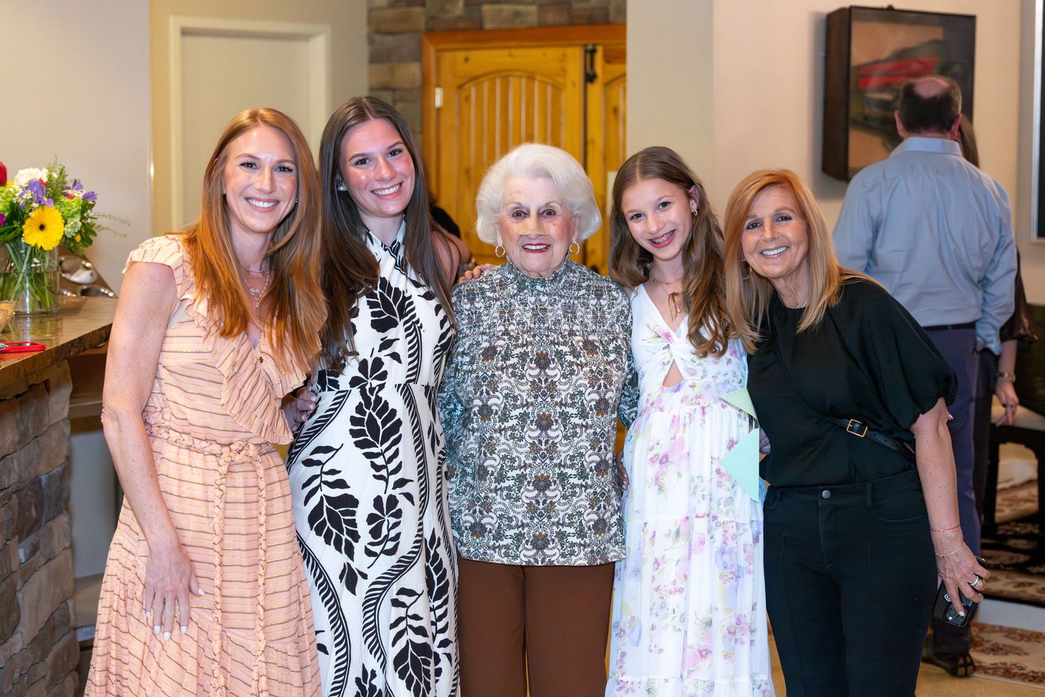 Five women of different generations smile together at an indoor family gathering celebration.