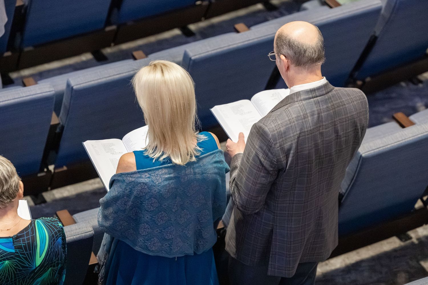 Two people standing in auditorium seating, reading from open books or programs during an event.