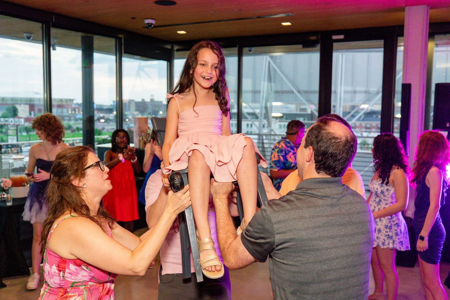 Young girl lifted by two adults at a festive indoor party with colorful lighting and guests in background.