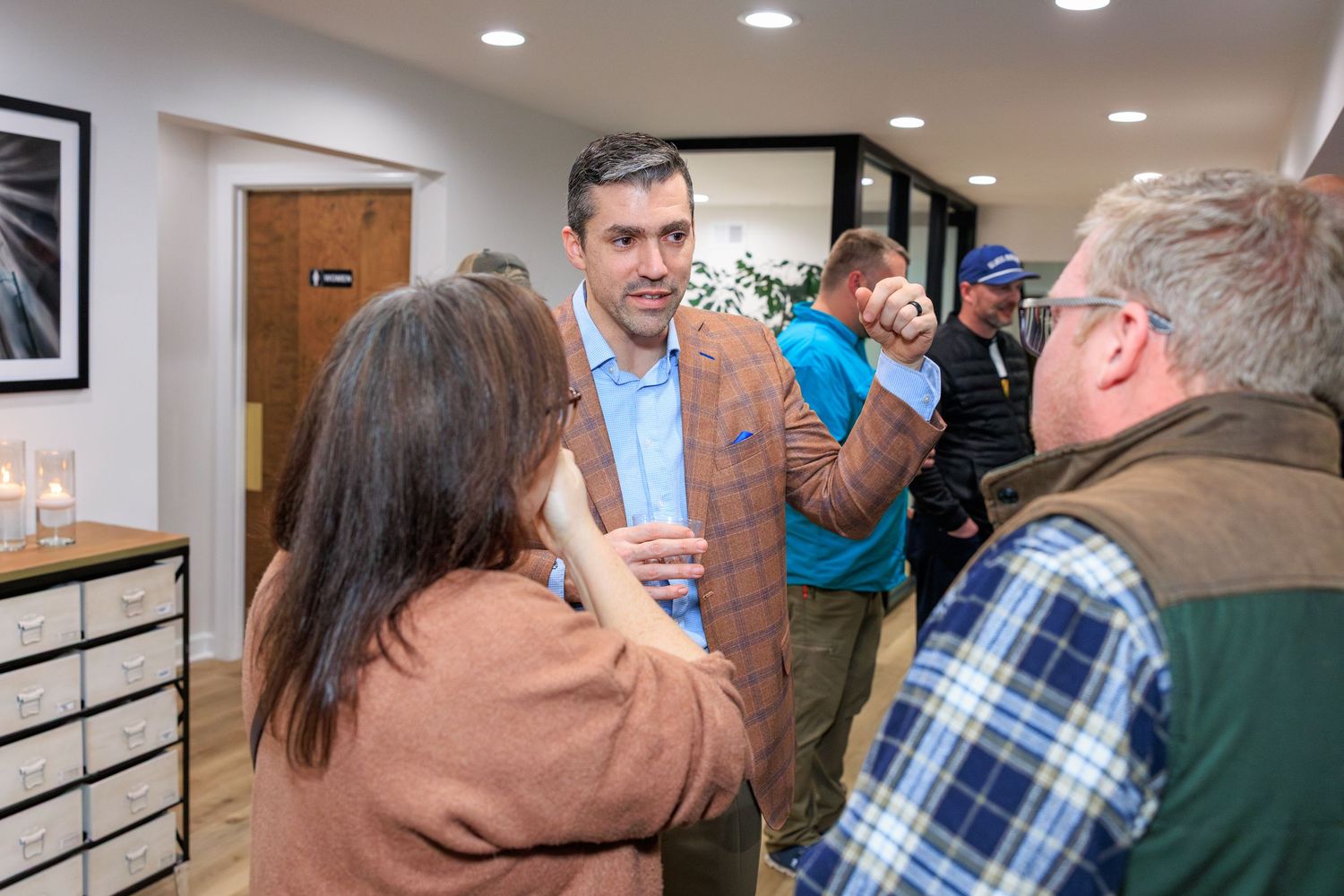 A group of people gathered in an office hallway having a conversation near a white wall with recessed lighting.