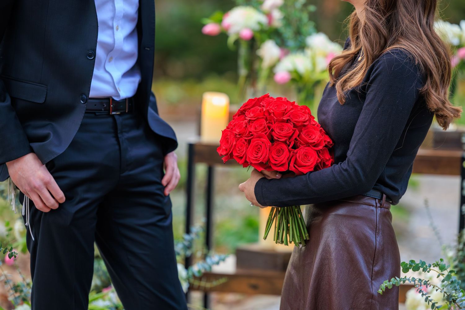 Wedding party members in formal attire stand together holding bouquets of red and white flowers in an outdoor garden setting.