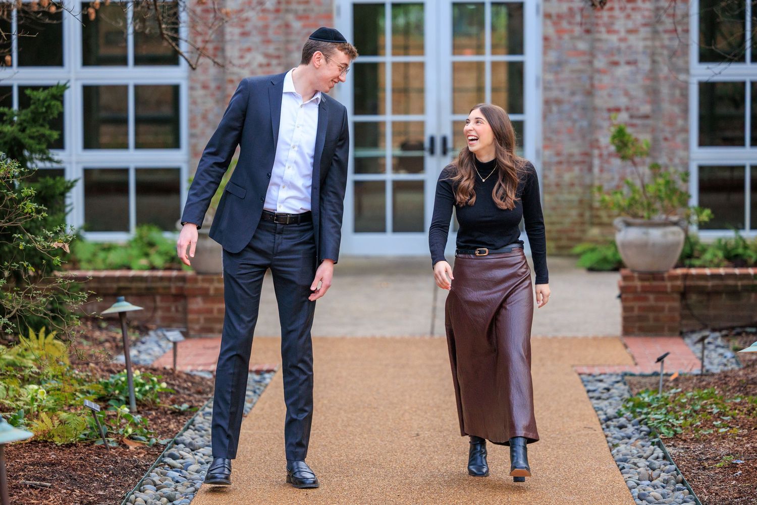Two people walk together on a paved pathway surrounded by landscaping and brick buildings.