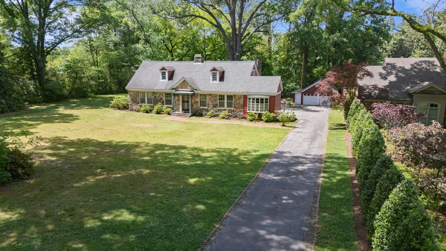 Charming brick Cape Cod home with manicured lawn, hedge-lined driveway, and mature shade trees.