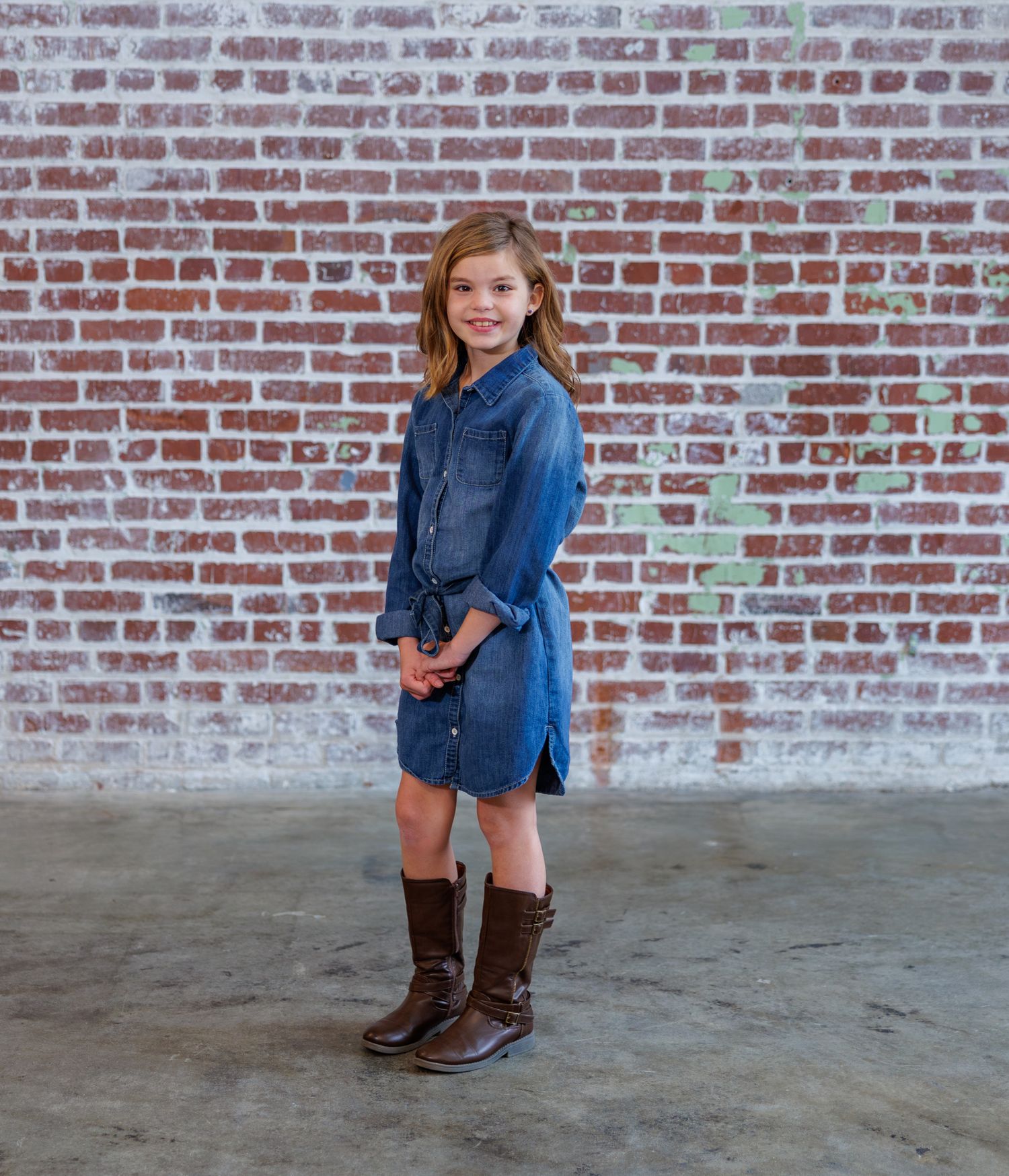 A child in a denim jacket and brown boots stands against a rustic brick wall background.