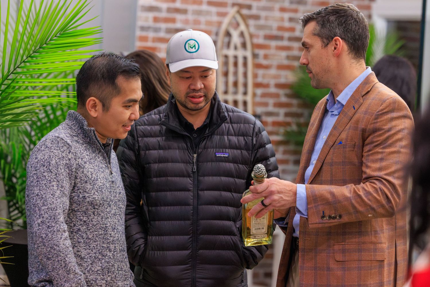 A group of men in casual attire gather together outdoors near a brick wall with palm fronds visible in the background.