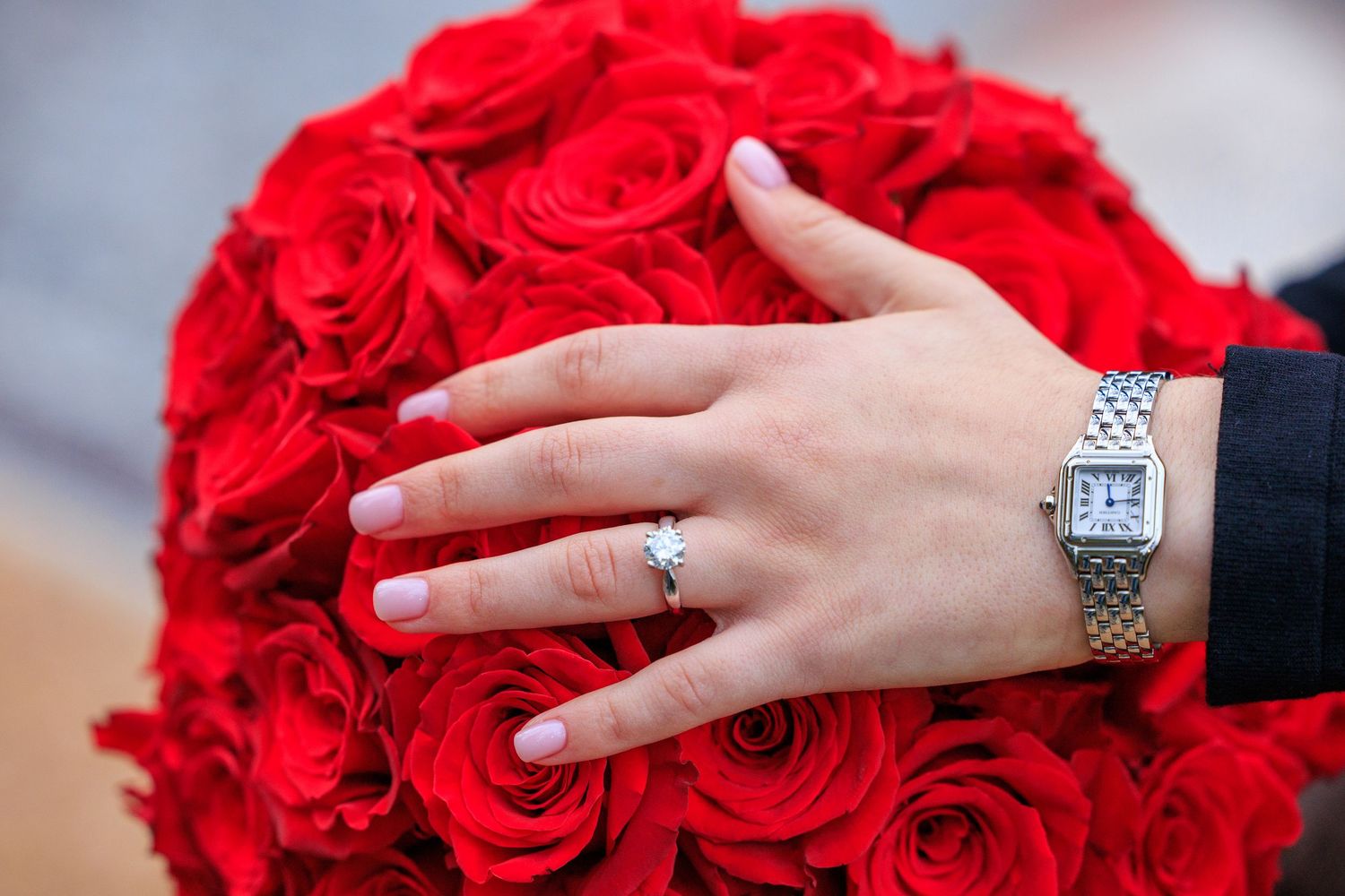A hand wearing a silver watch and diamond ring rests on a bouquet of vibrant red roses.