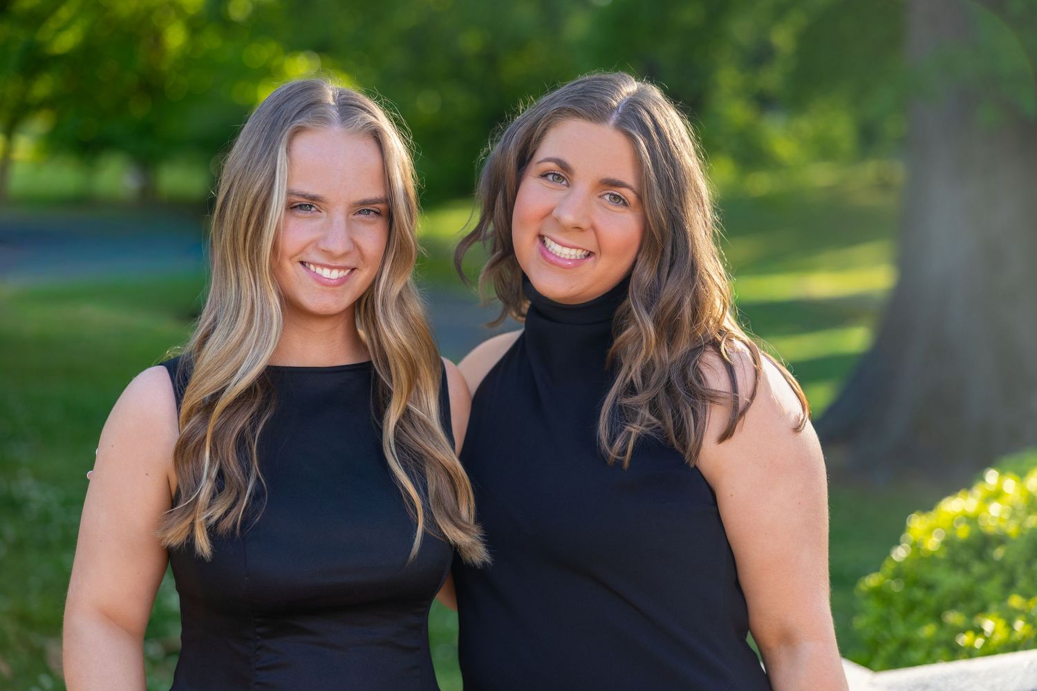 Two smiling women in black outfits pose outdoors in a lush green park setting on a sunny day.