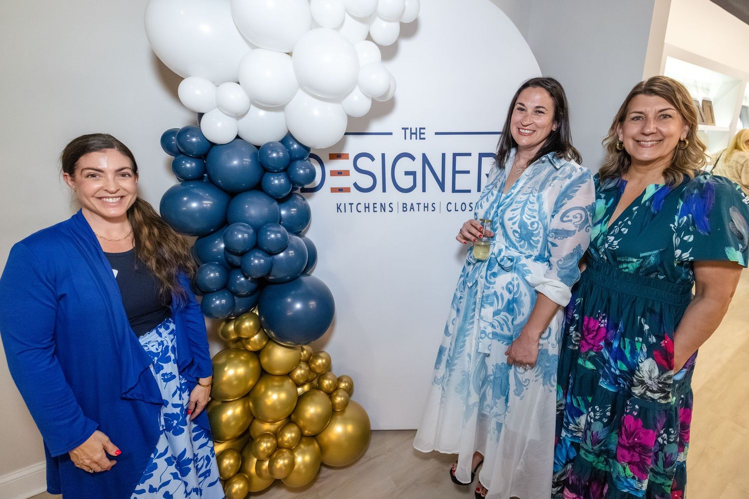 Three women smiling at The Designer Kitchens Baths Closets showroom opening event with balloon display.