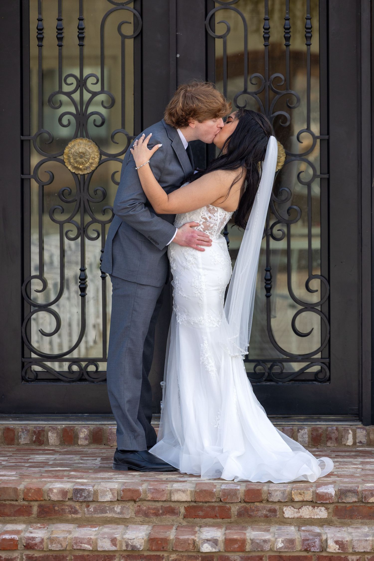 A couple in formal wedding attire share a romantic kiss on brick steps in front of an ornate iron gate.