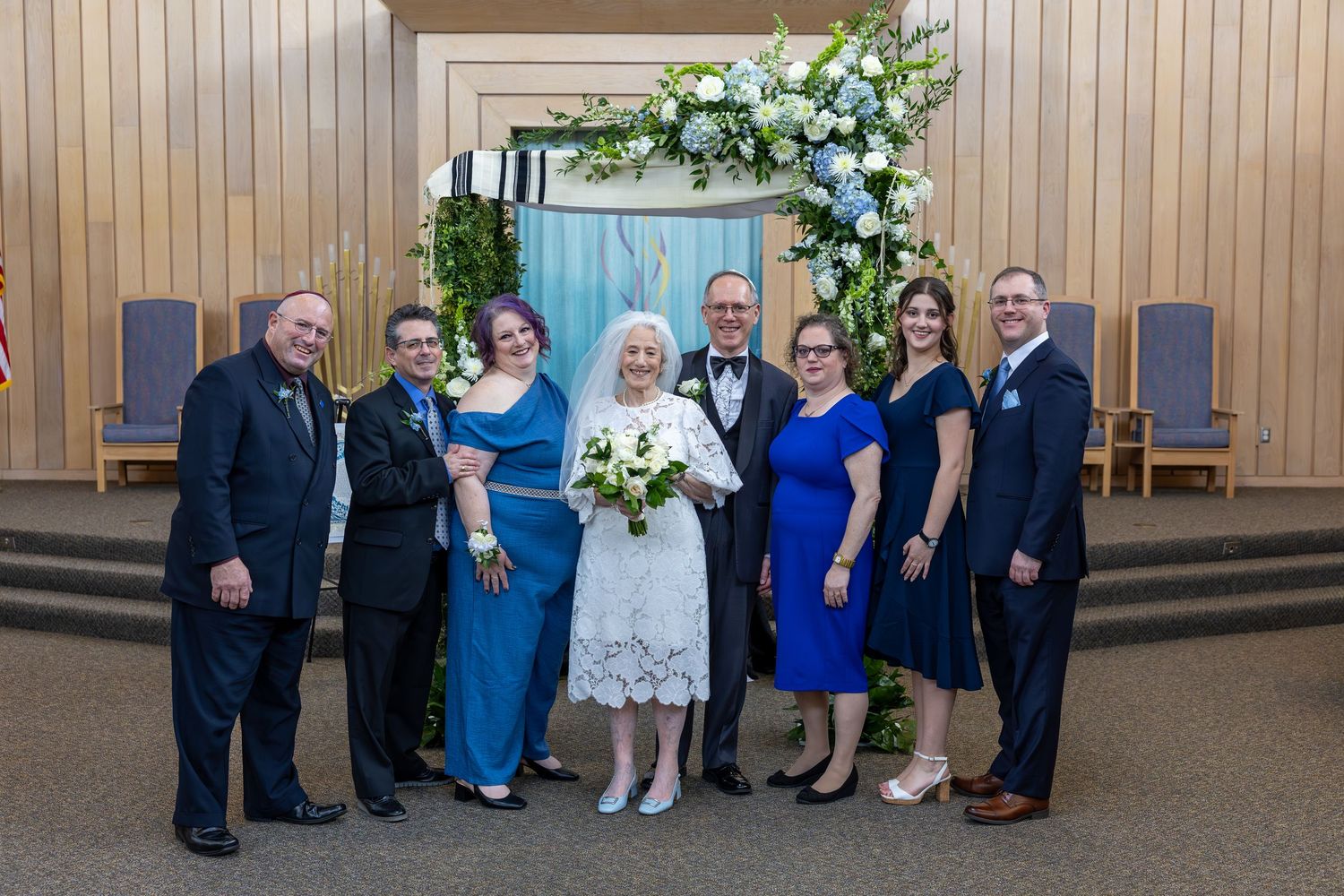 A group of formally dressed guests gather in a hallway with wooden paneled walls and floral decorations.