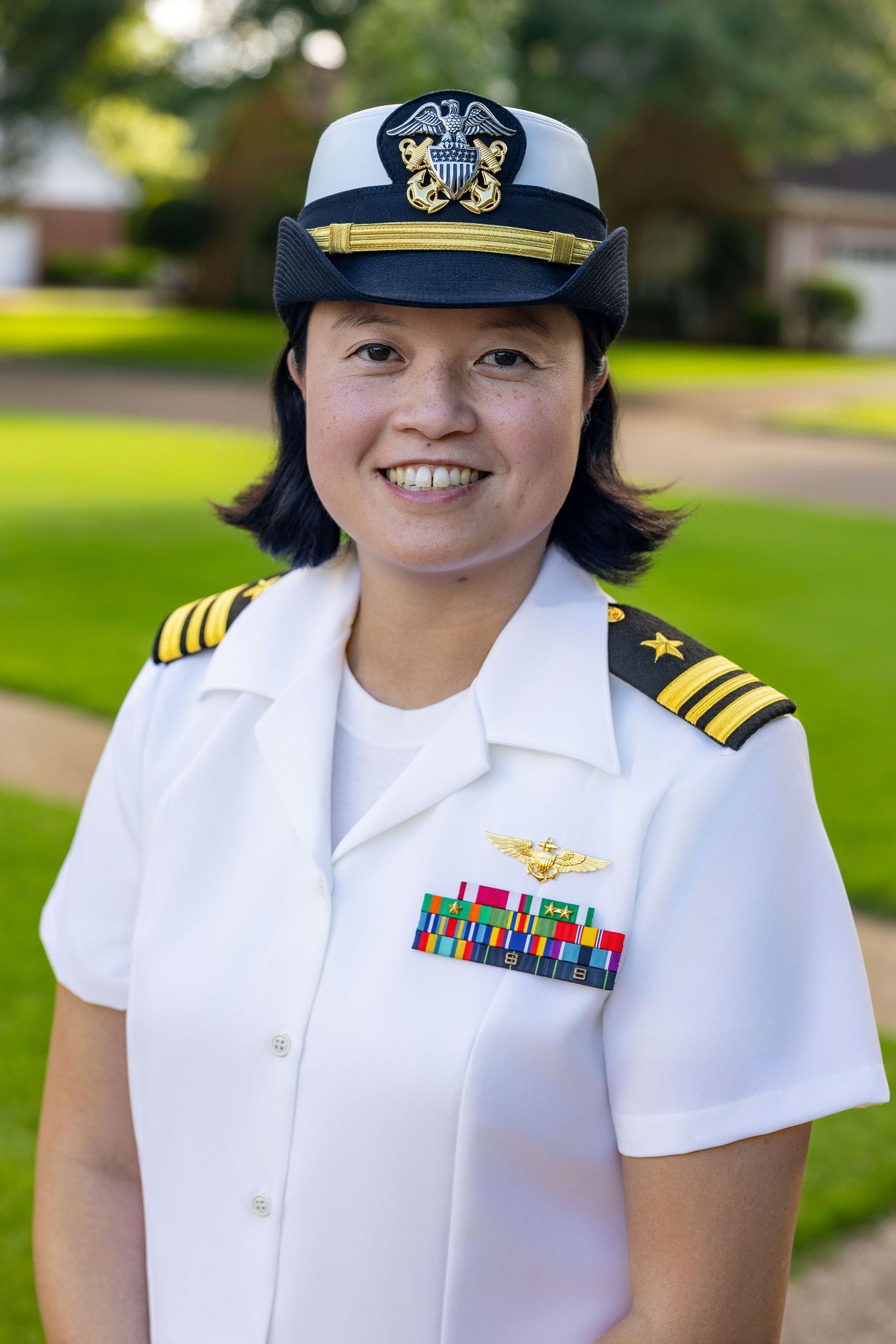 Female U.S. Navy officer in white dress uniform with gold insignia, ribbons, and officer's cap, smiling outdoors.