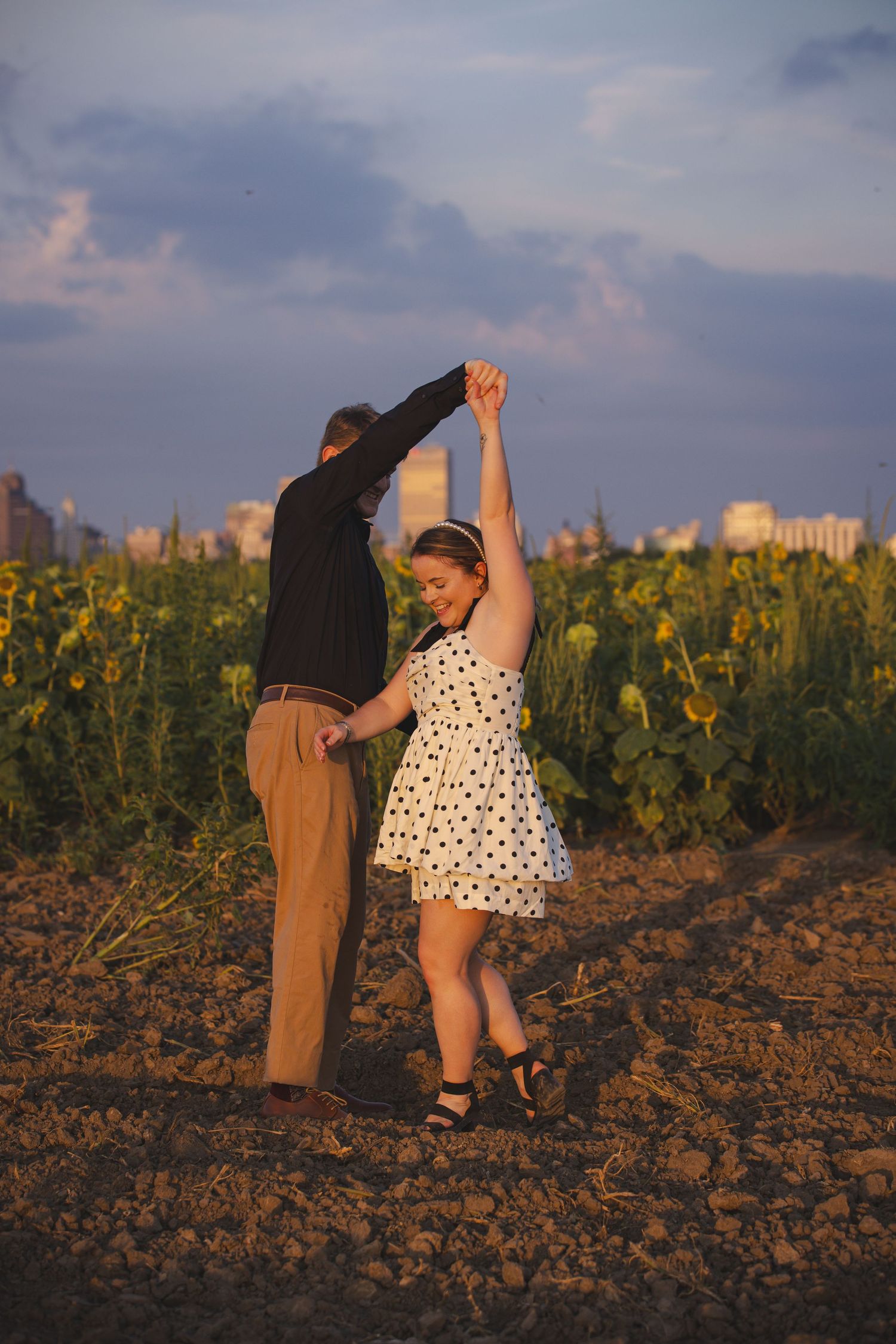 Couple dancing in a sunflower field at sunset with city skyline in background.