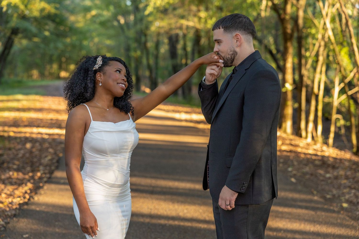 A couple in wedding attire walks along a tree-lined path during golden hour.
