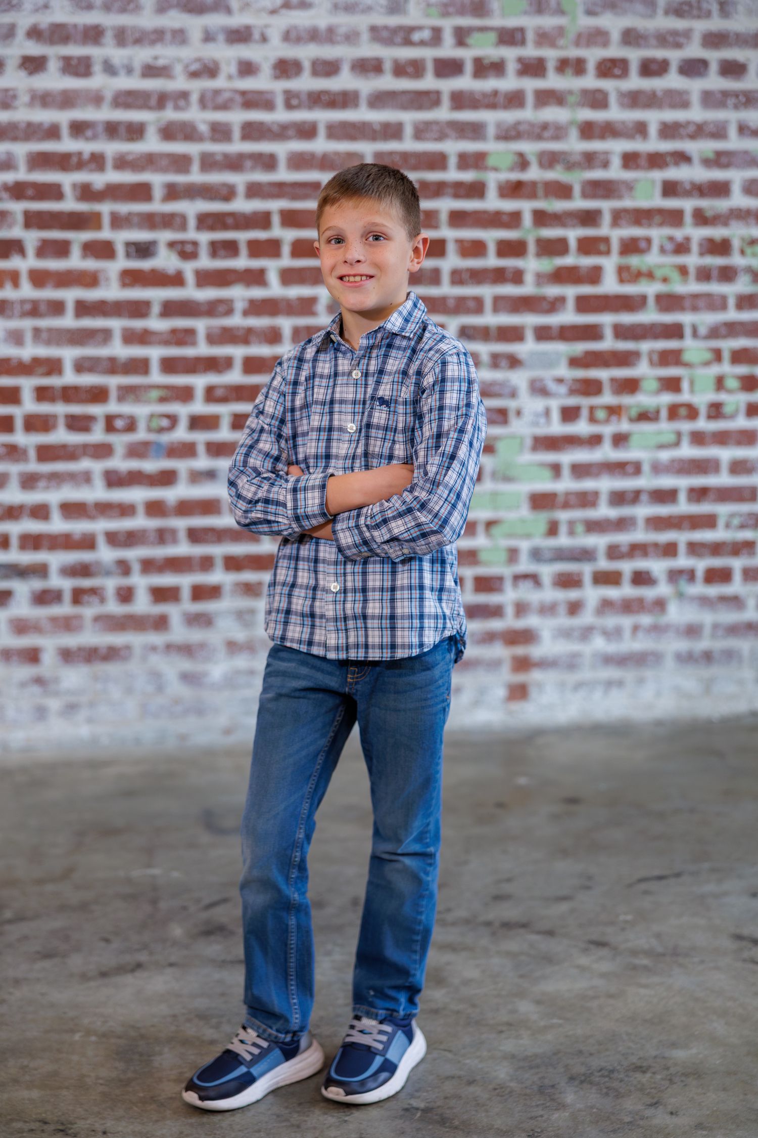 A young figure poses playfully against a brick wall wearing a plaid shirt and jeans.