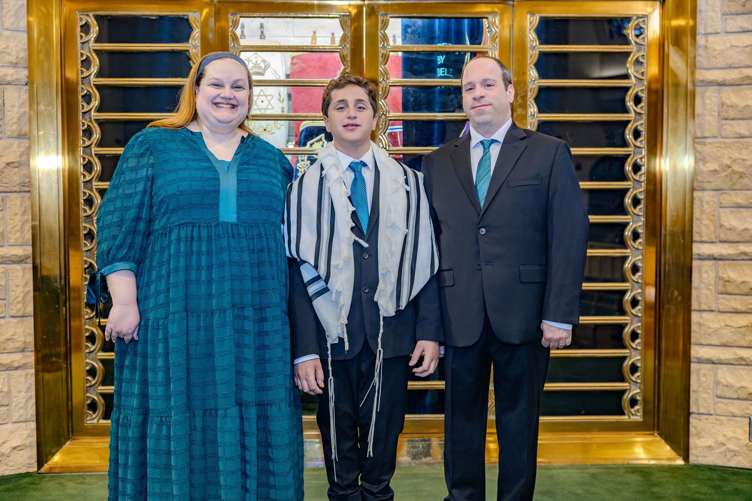 Three people stand together in formal attire in front of wooden wall panels at an event.