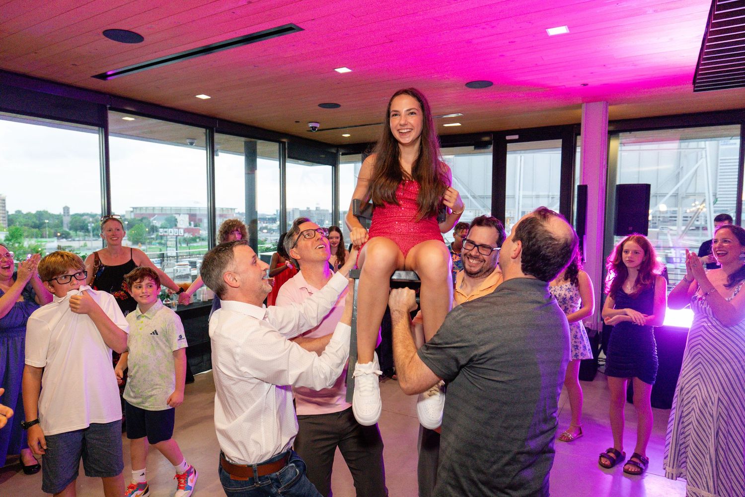 Girl in red dress lifted by guests at a festive Bat Mitzvah celebration in a modern venue.