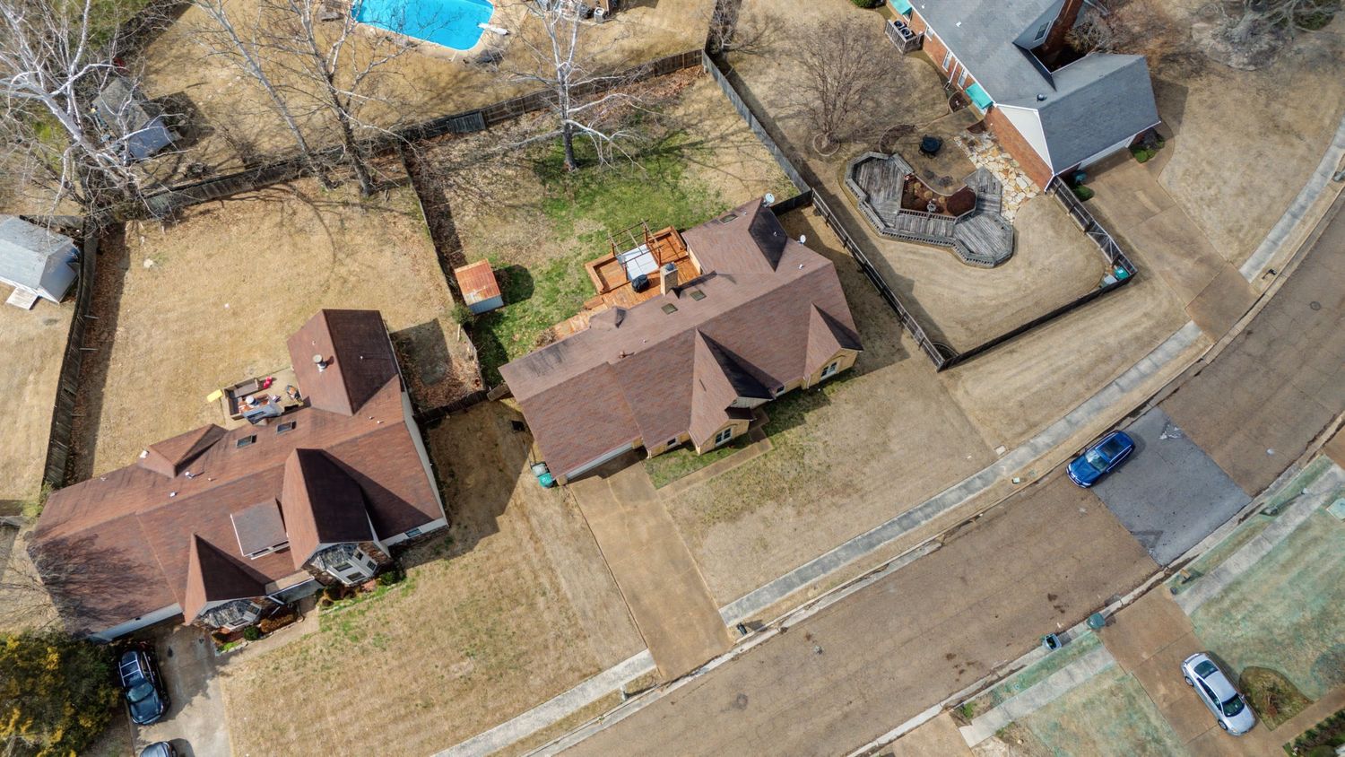 Aerial view of three brown-roofed houses with a dirt road and parked cars in a rural setting.