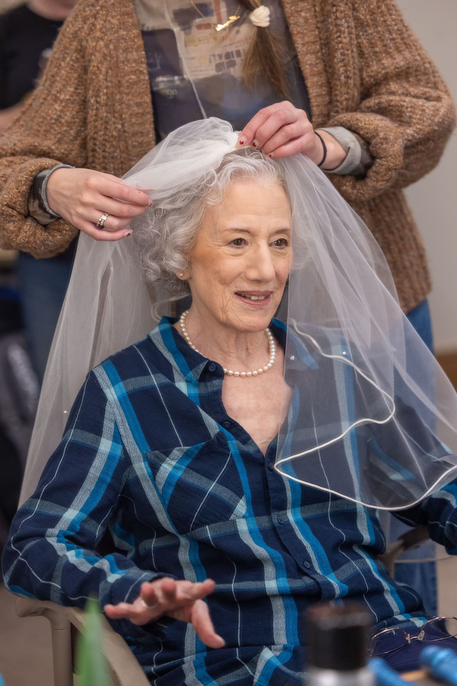 A person wearing a blue plaid shirt has a white veil placed on their head while sitting in a chair.