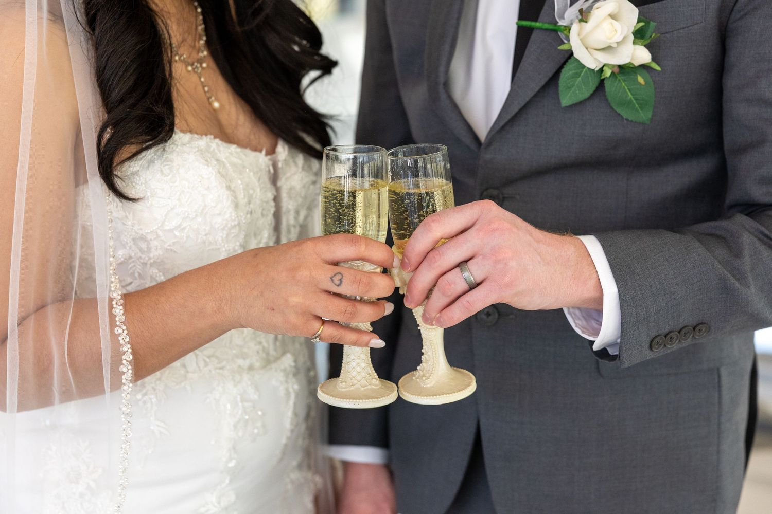 Newlyweds toast with champagne flutes, showing lace wedding dress and gray suit with white rose boutonniere.