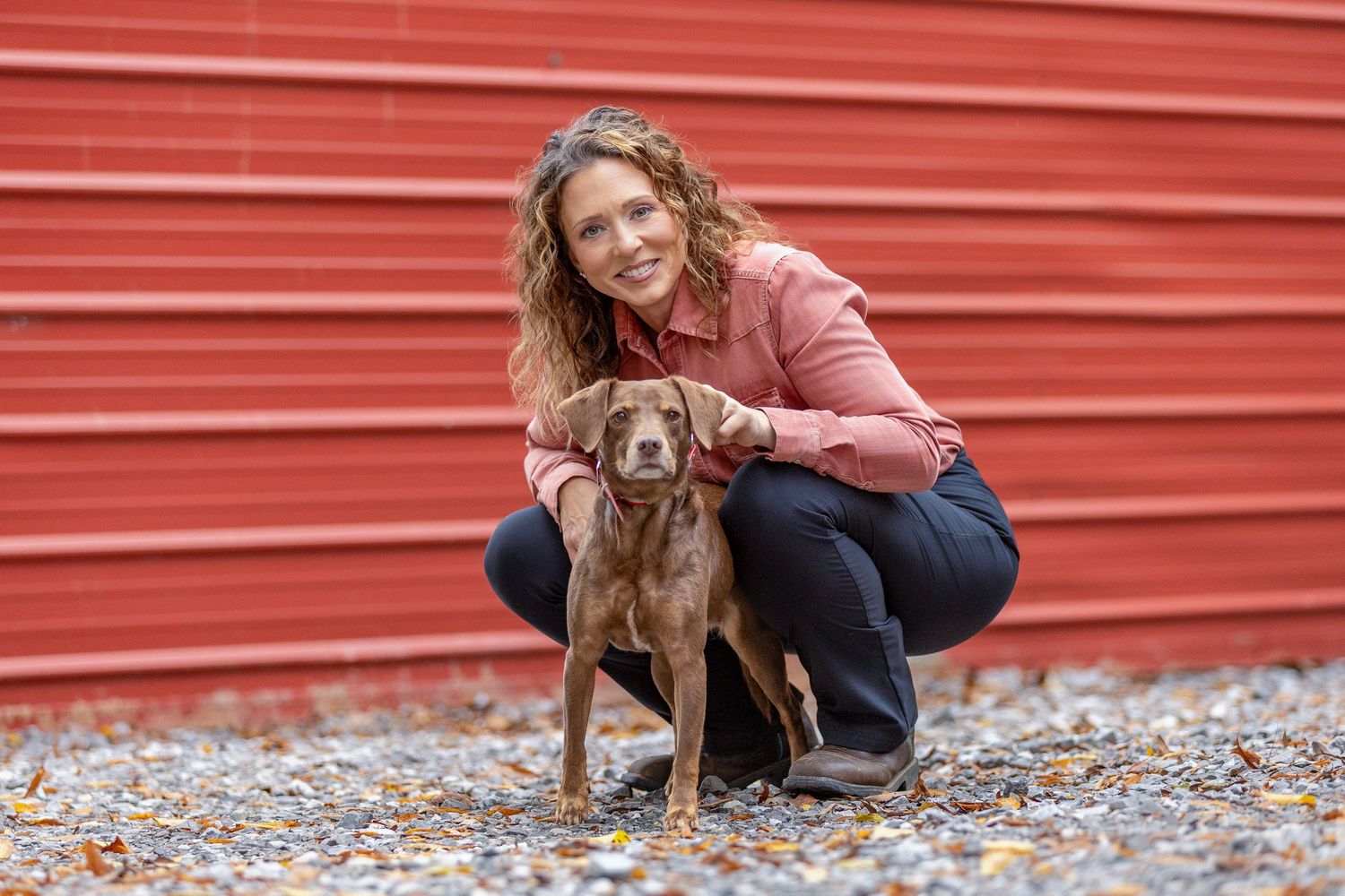 A person in pink jacket and jeans poses with two black dogs against a red wall.