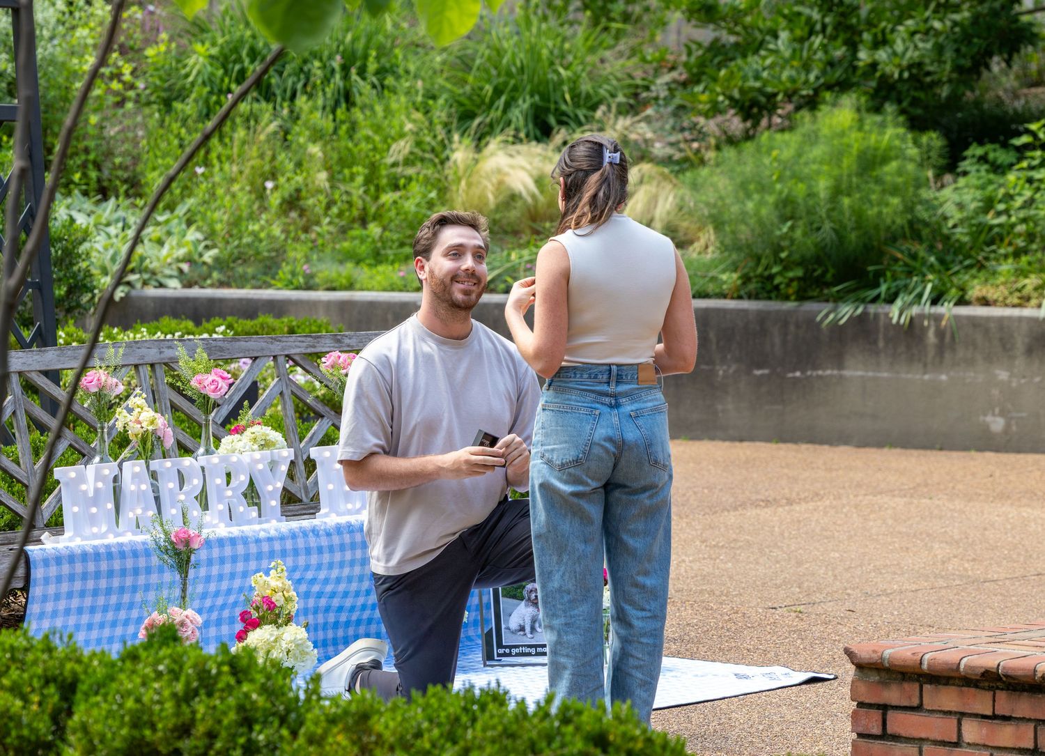 Man kneeling on one knee proposing to woman outdoors near floral decorations in a lush garden setting.