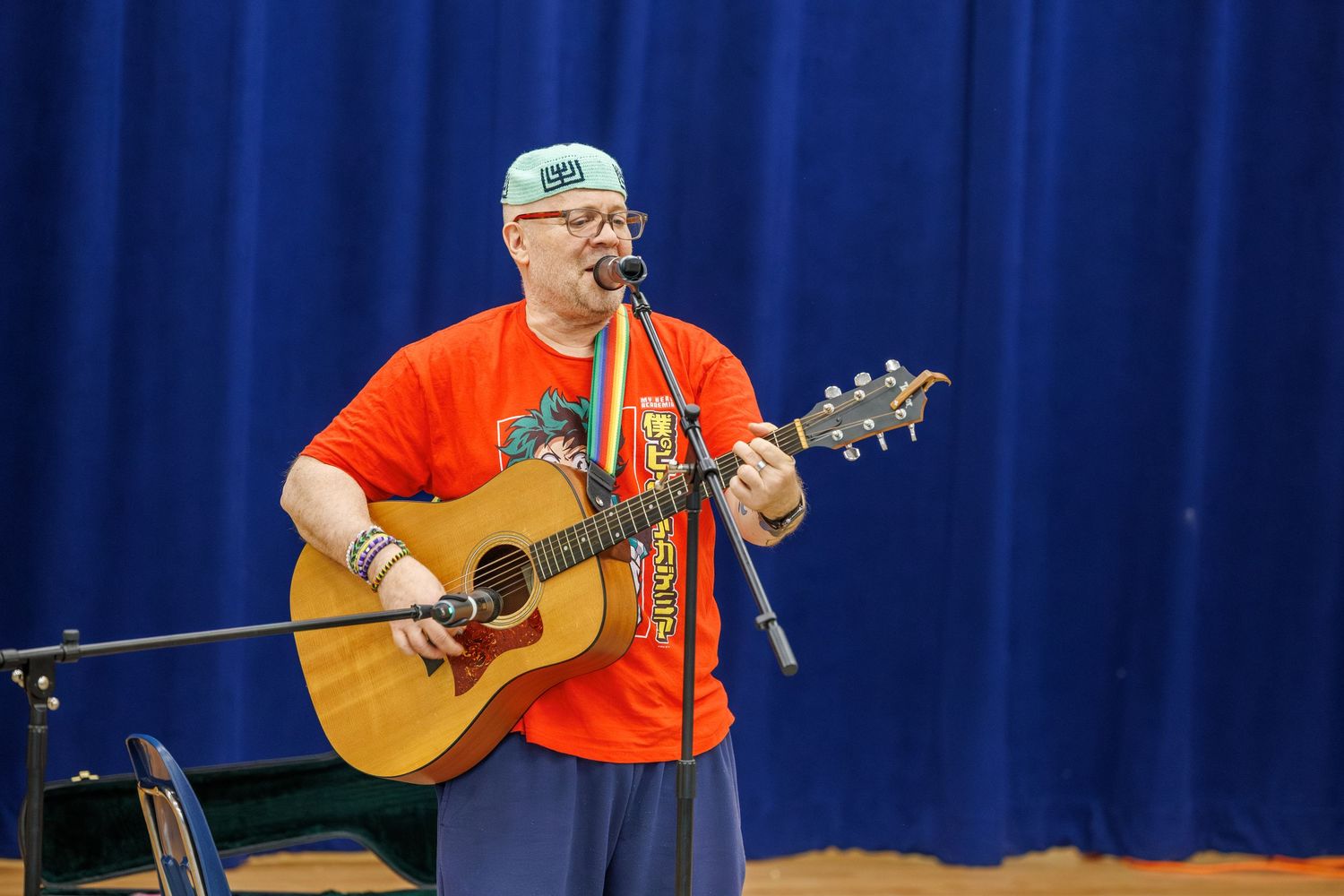 A musician performs on stage with an acoustic guitar wearing an orange t-shirt and baseball cap against a blue curtain backdrop.