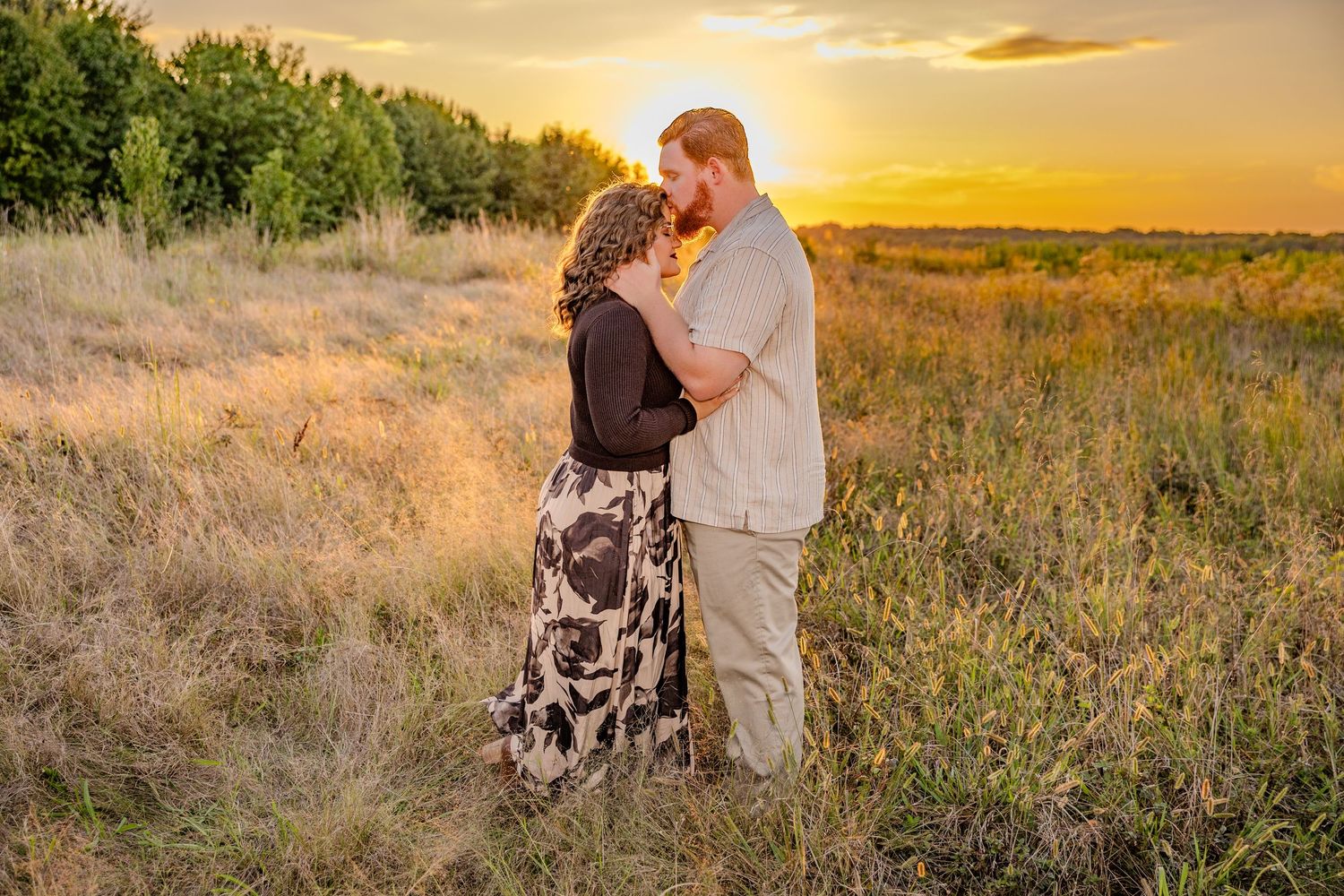 Loving couple poses in field with dramatic sunset sky behind them.