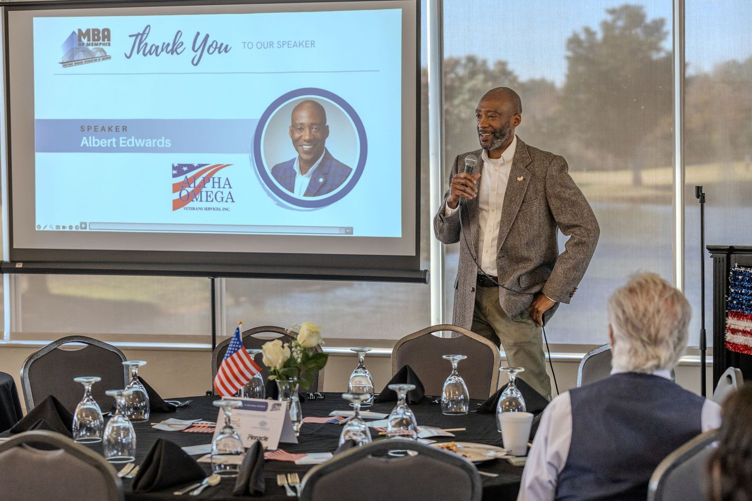 Speaker presenting at podium with projection screen displaying presentation in background.