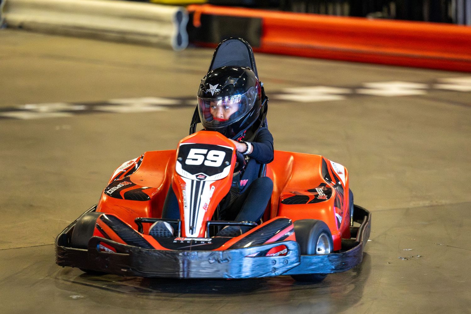 A go-kart racer in blue helmet drives an orange racing kart on an indoor track.