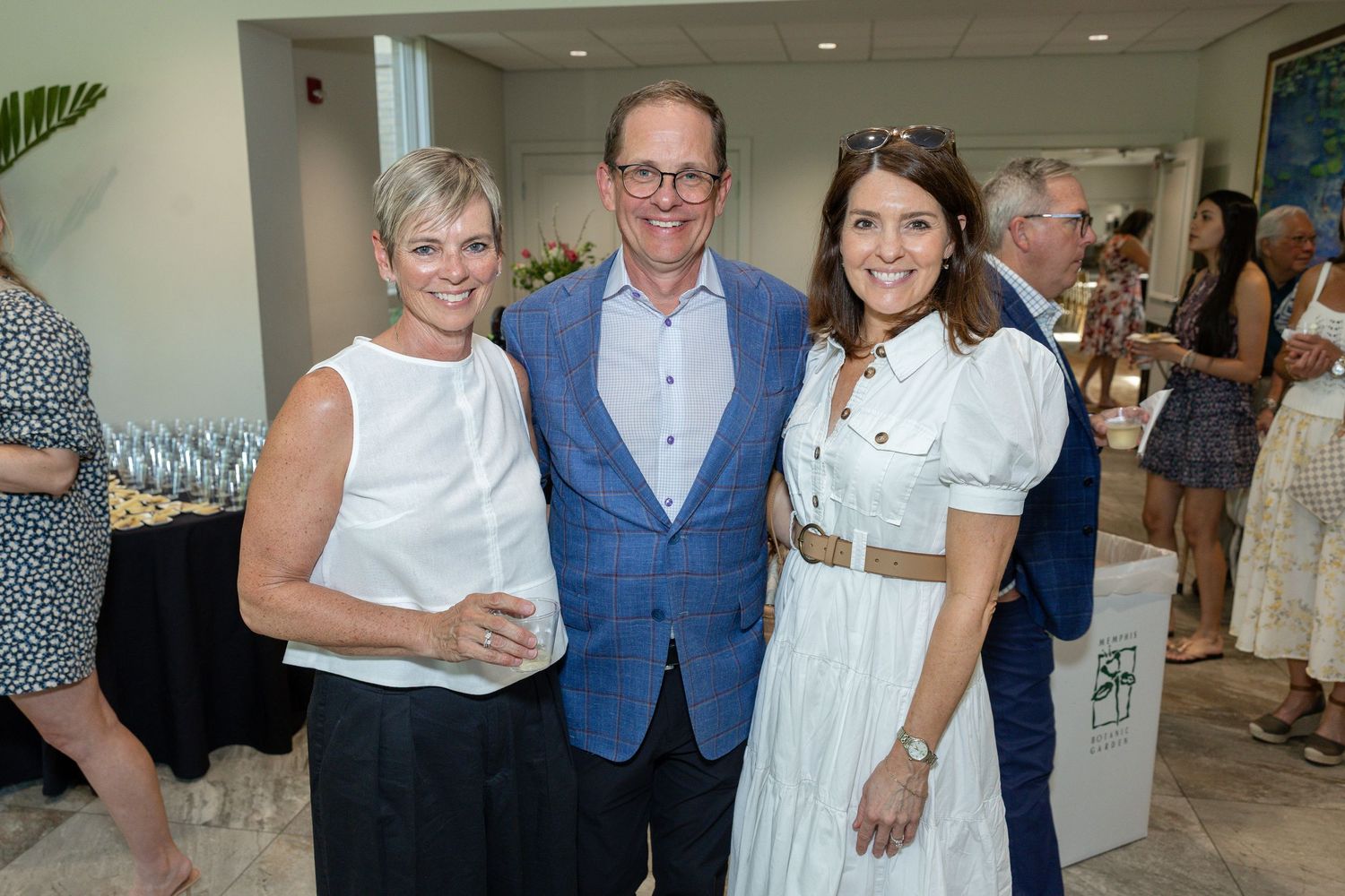 Three smiling guests pose together at an elegant indoor social event, dressed in smart casual attire.