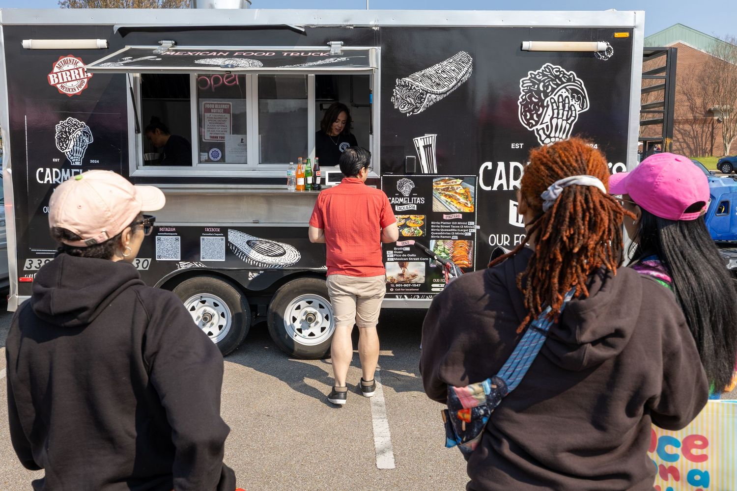 A line of customers wait outside a food truck in a parking lot on a sunny day.