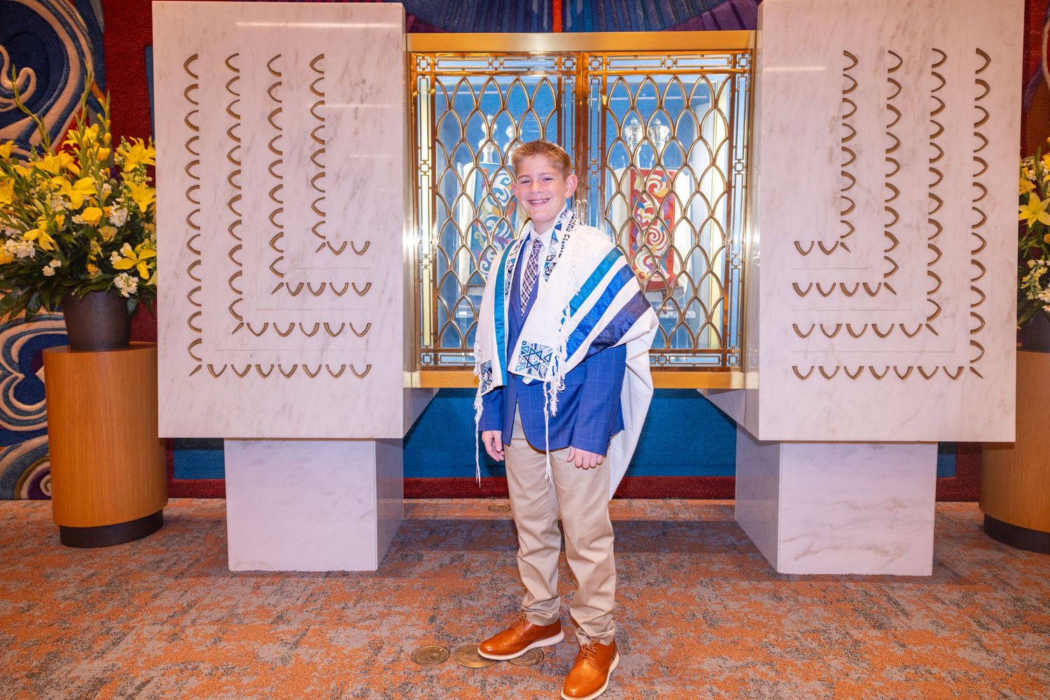 Young boy in Bar Mitzvah attire wearing a blue and white tallit, posing proudly in a decorated synagogue.