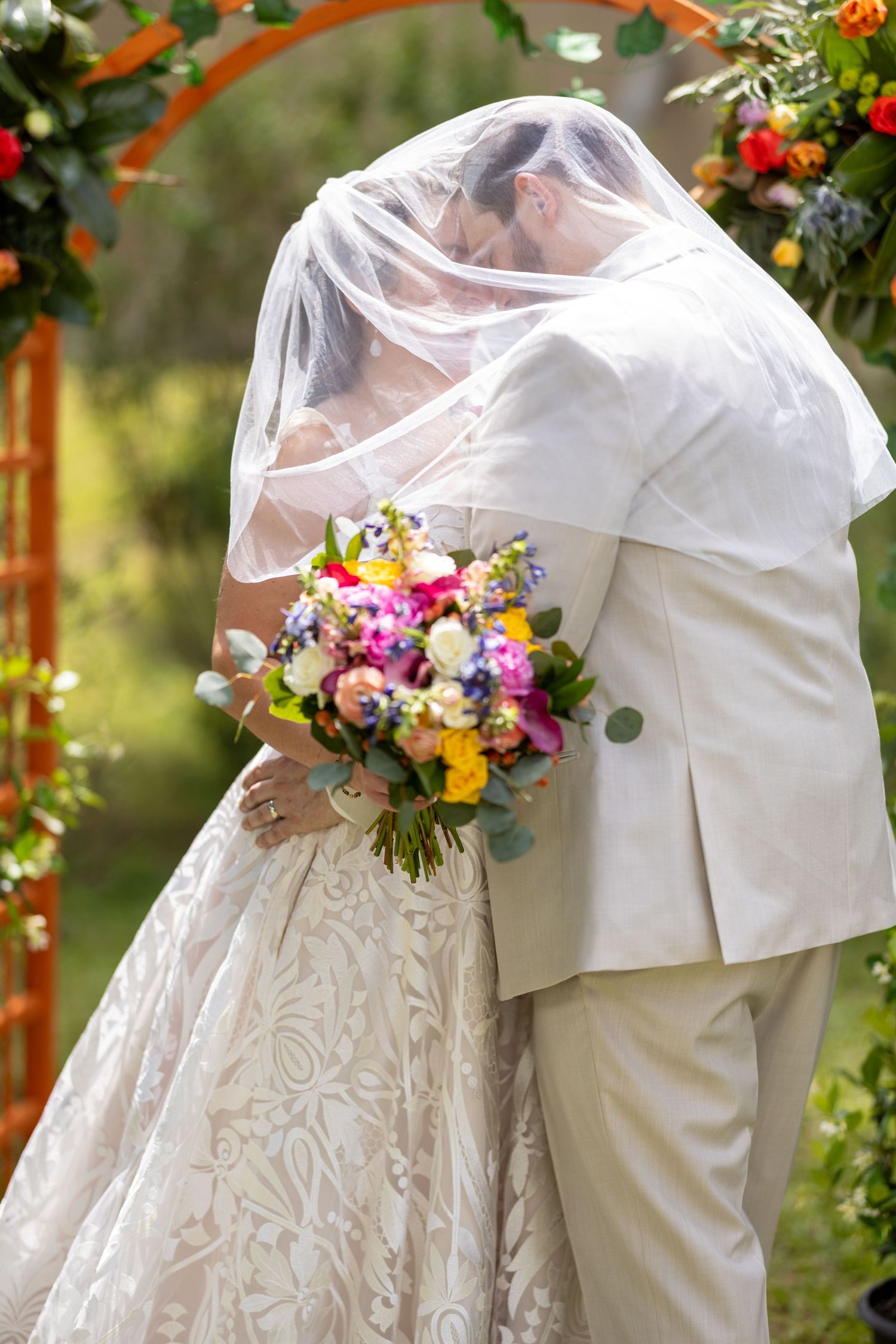 Bride and groom sharing a kiss under a veil, holding a colorful bouquet at an outdoor wedding ceremony.