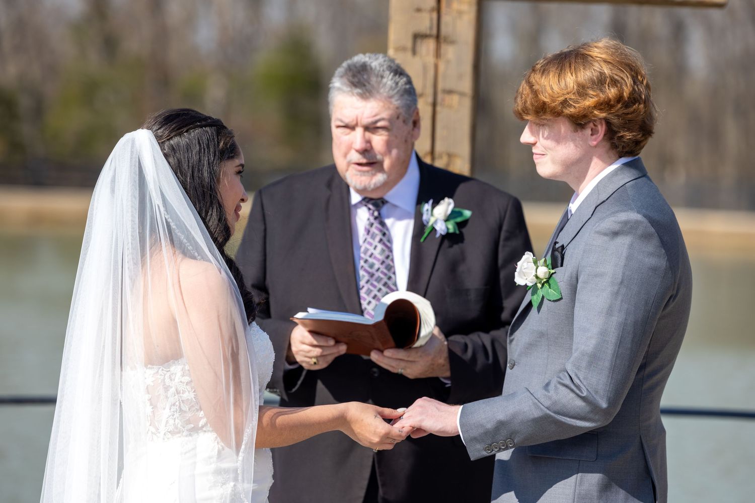An outdoor wedding ceremony with a couple and officiant standing together on a sunny day by a lake.