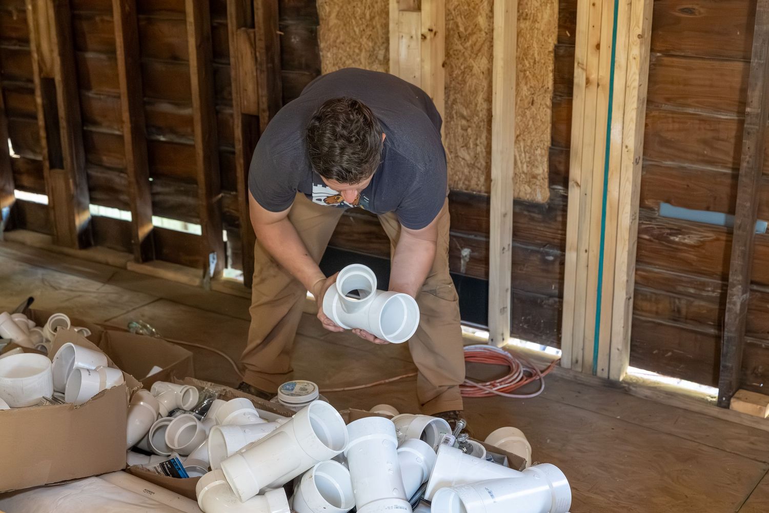 Plumber sorting PVC pipe fittings on the floor of a wood-framed building under construction.