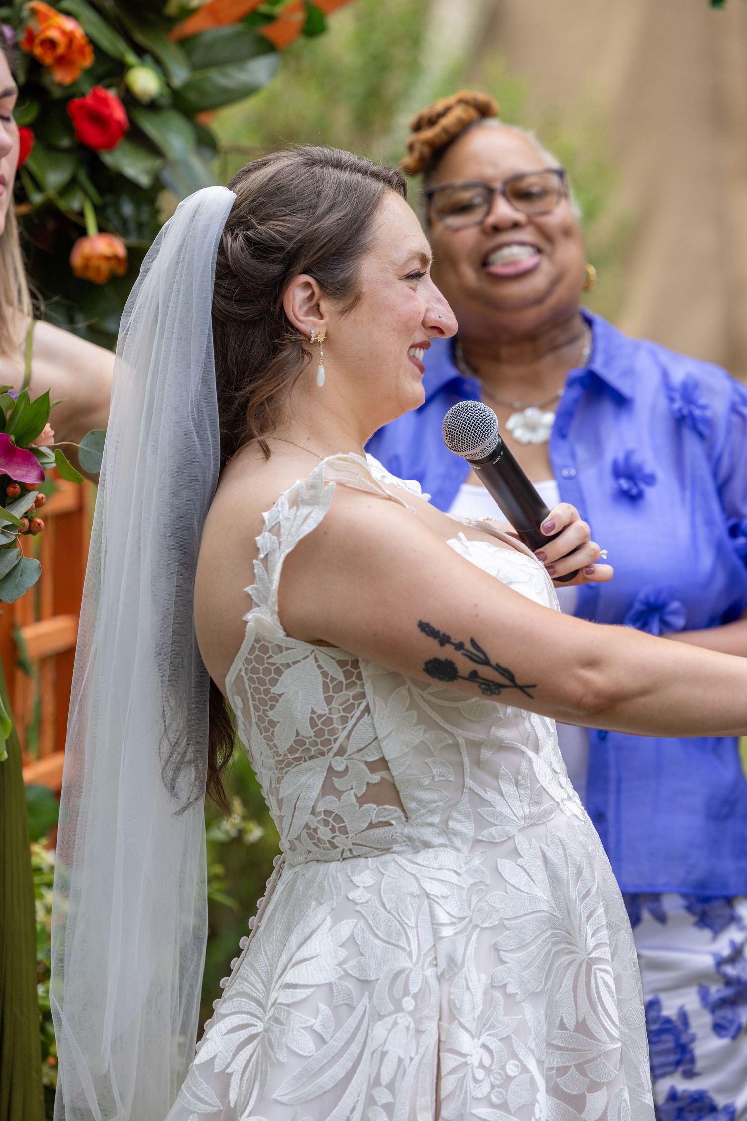 Bride in lace wedding dress and veil laughs joyfully during ceremony while officiant smiles holding microphone.