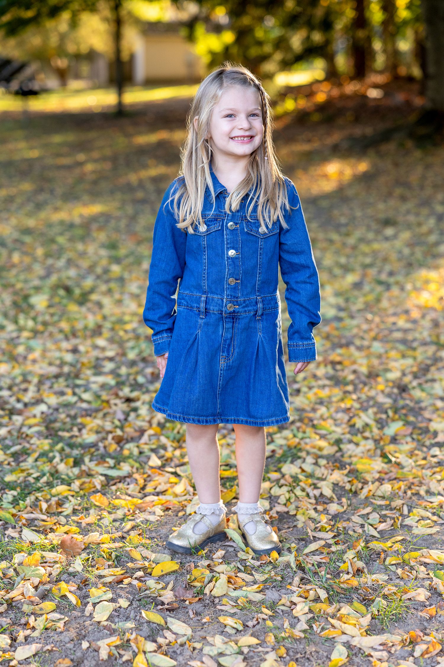 Child wearing blue denim dress standing on autumn leaves.