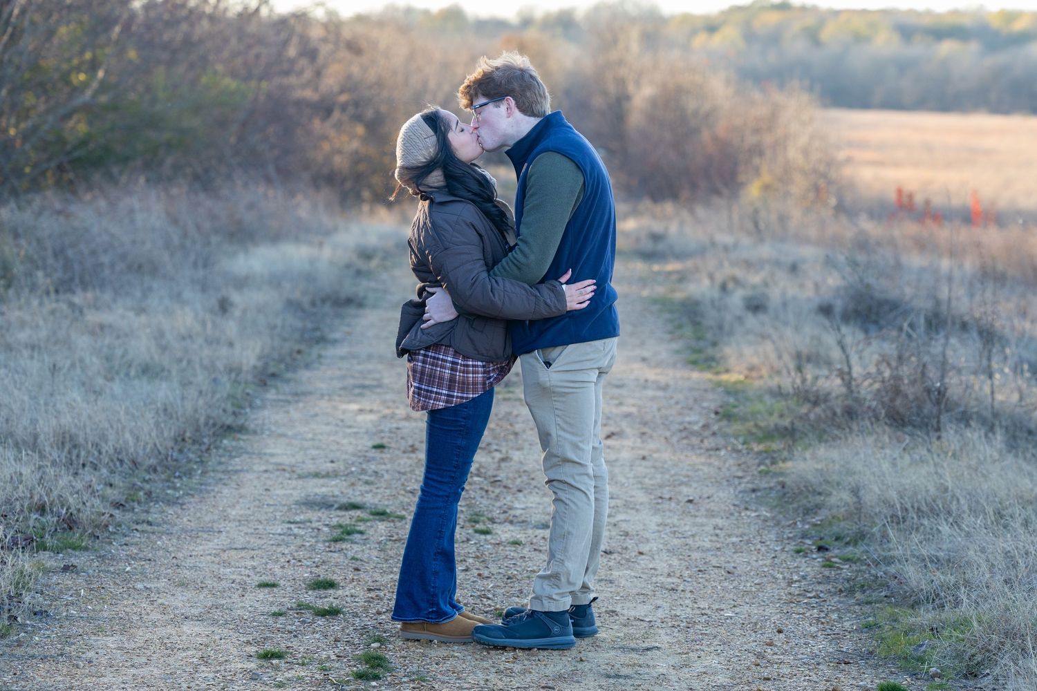 A couple embraces on a dirt path during sunset in a golden field.