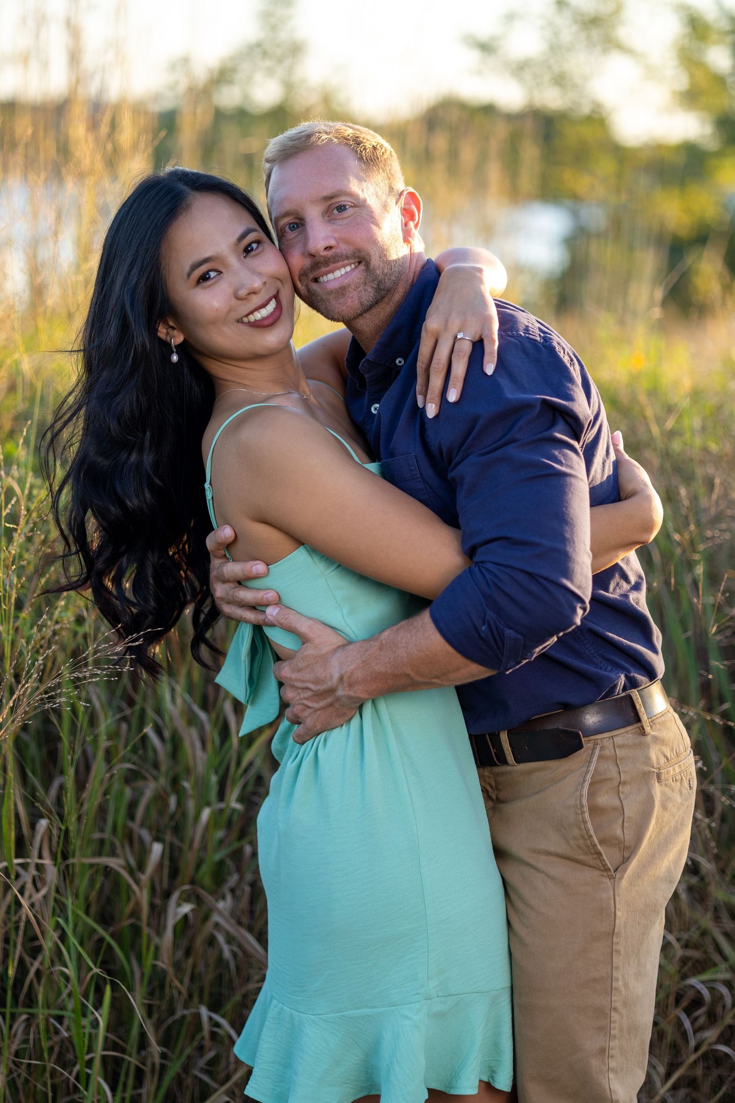 A couple embraces in a field during golden hour, with the woman wearing a mint green dress and man in navy blue shirt.