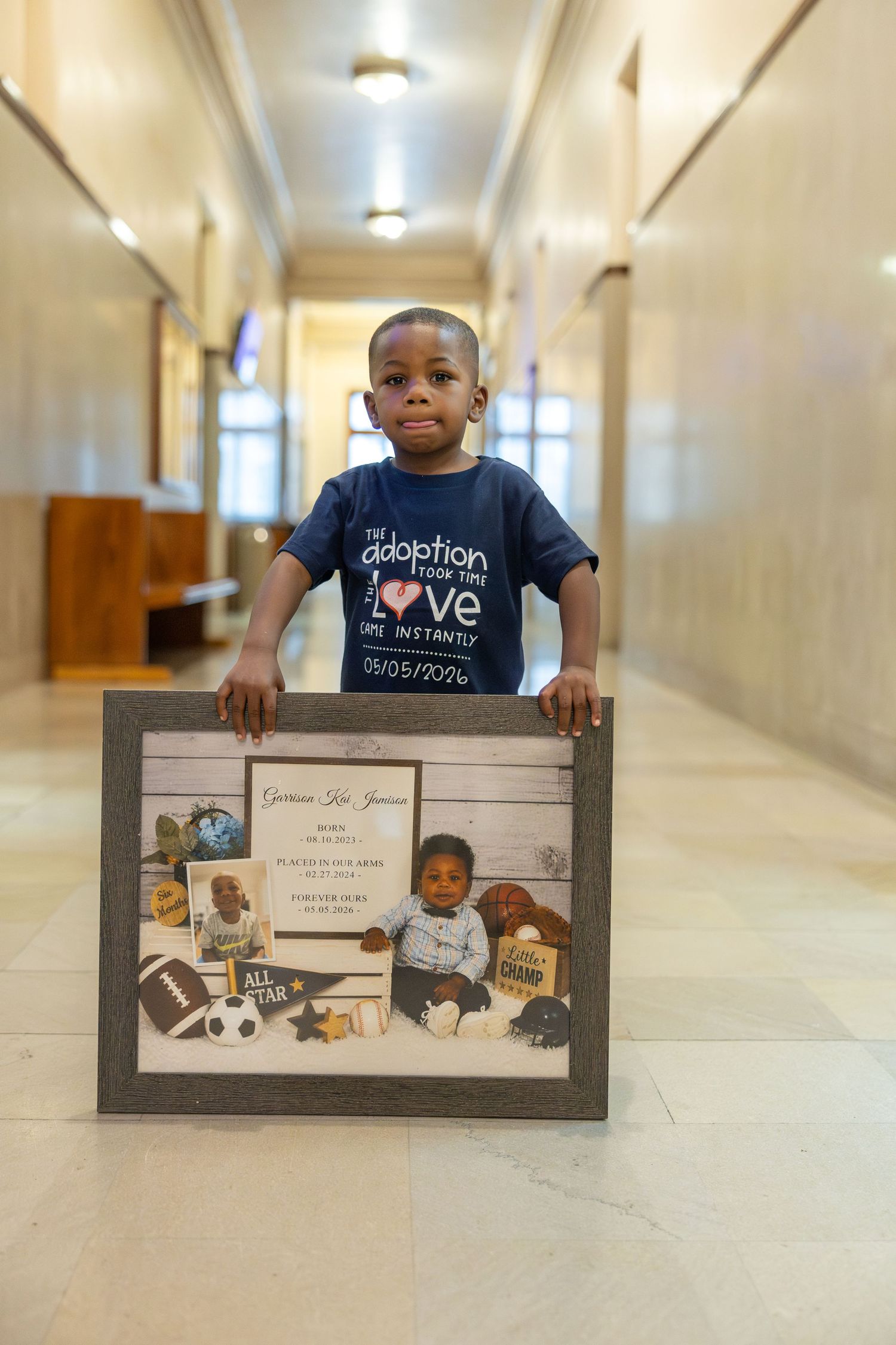 Young boy in adoption t-shirt proudly holds a framed photo collage in a courthouse hallway on his adoption day.