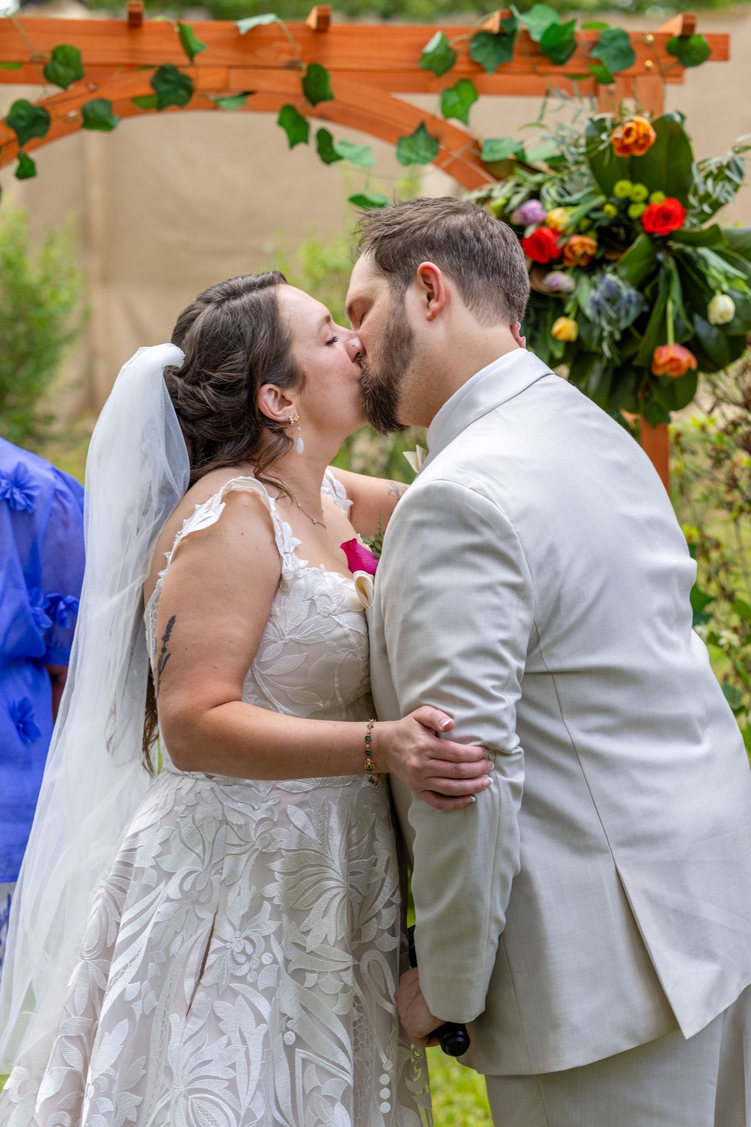 Bride and groom share their first kiss at outdoor wedding ceremony beneath a floral arch.