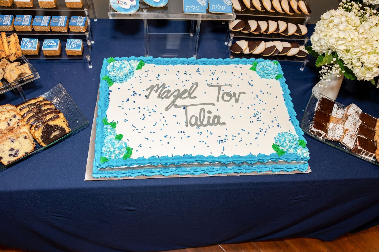 Mazel Tov Talia celebration cake with blue frosting and floral decorations on a navy blue tablecloth.