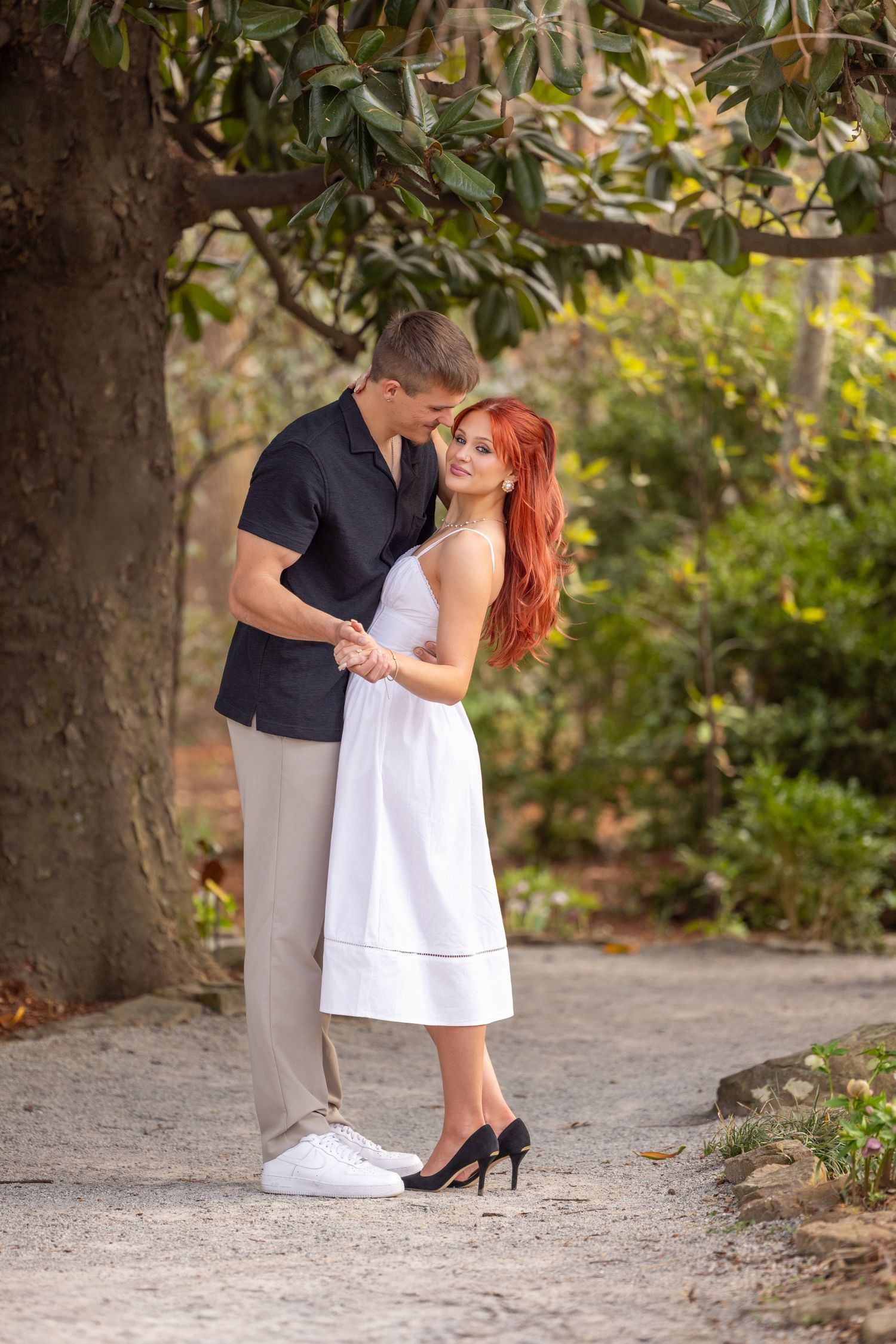 A couple shares an intimate dance moment on a path surrounded by trees and natural scenery.