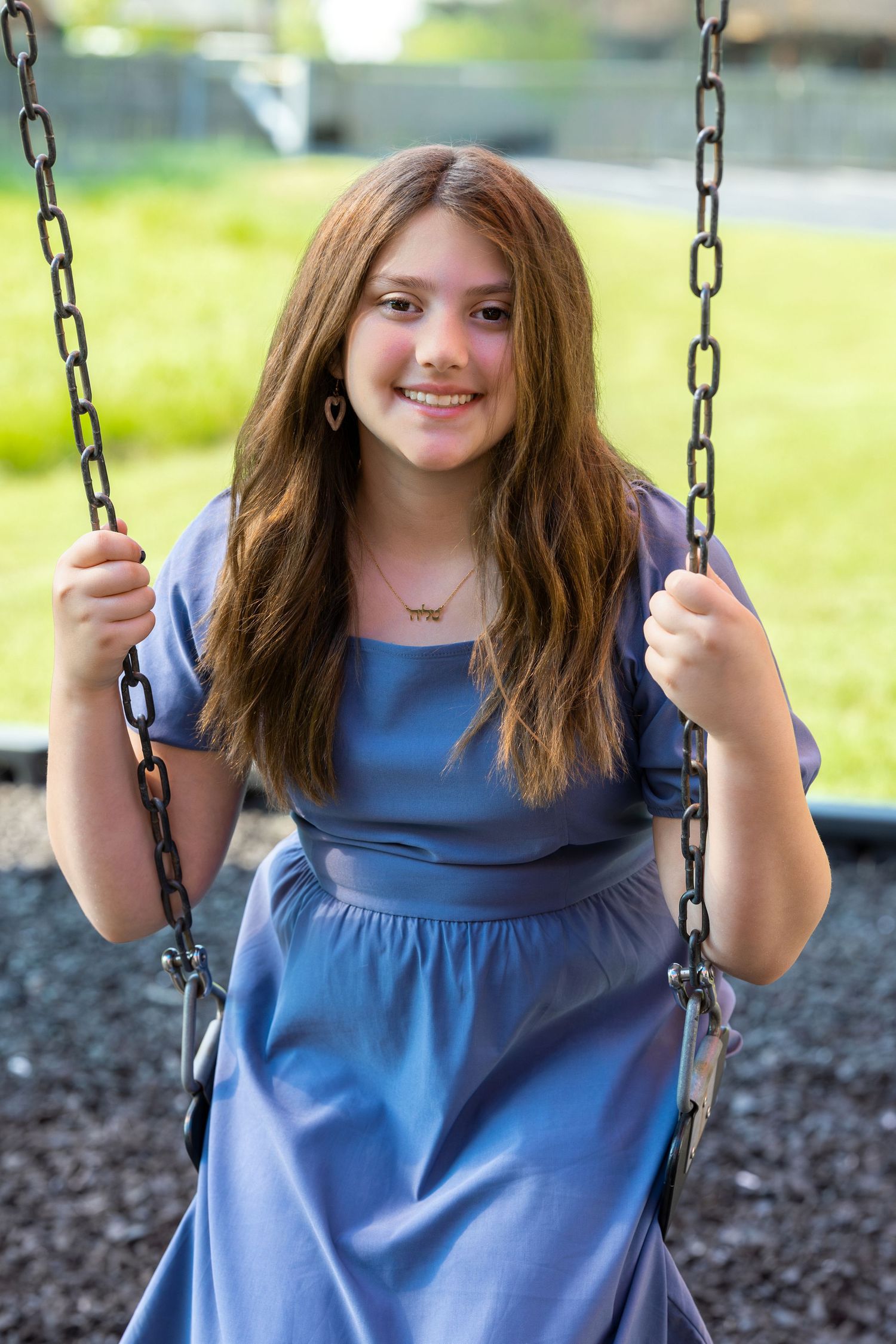 Smiling teenage girl in blue dress sitting on playground swing, holding chains, with green grass background.