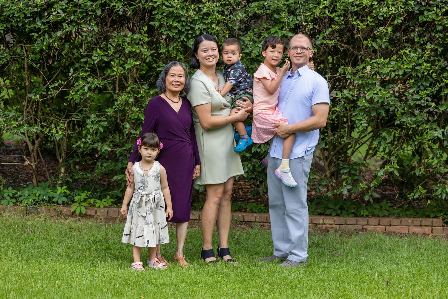 Family of six posing outdoors on green grass with lush ivy background, including grandparent, parents, and three young children.