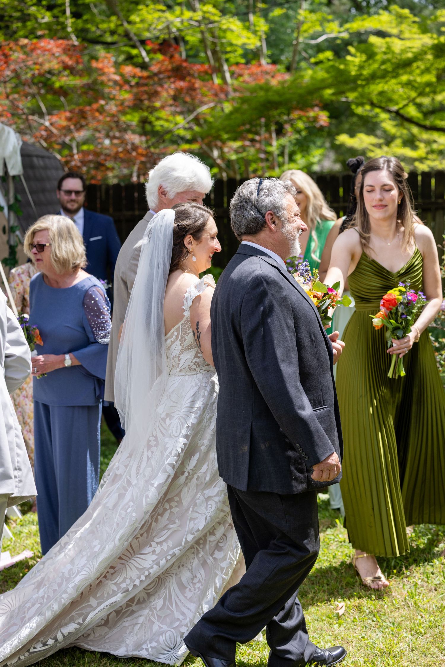 Bride in white lace gown walks with escort at outdoor wedding ceremony, bridesmaid in olive dress holds bouquet nearby.