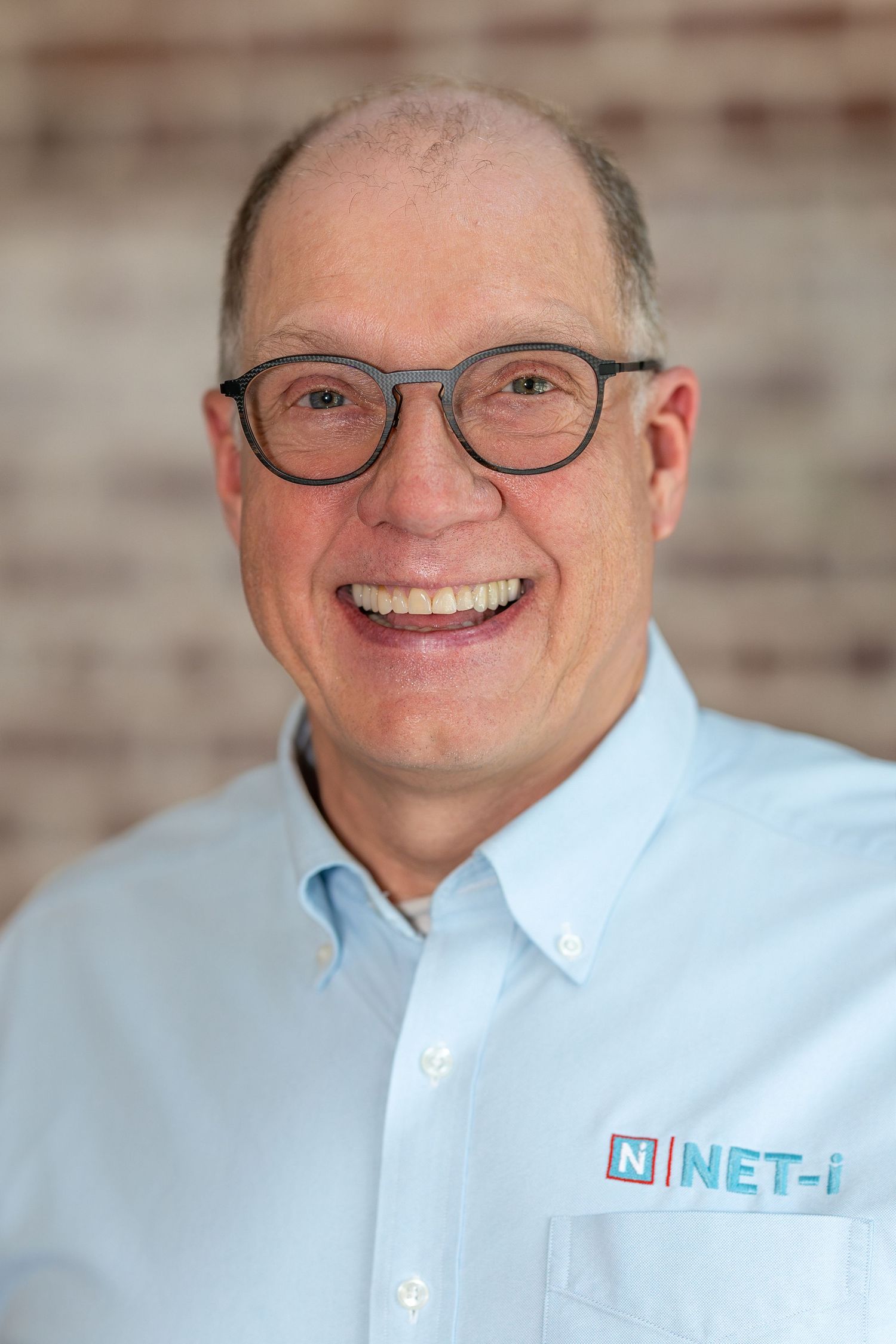 Smiling middle-aged man wearing glasses and a light blue NET-i branded shirt, posed against a brick wall background.