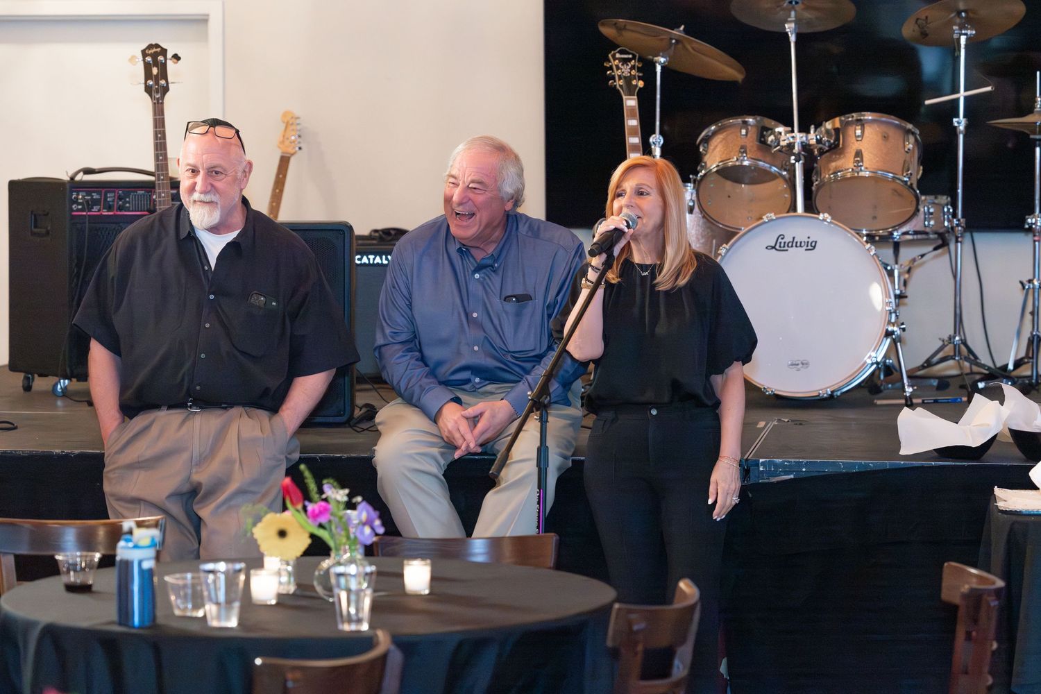 Three people smiling on stage near a microphone with drums and amplifiers in a music venue.