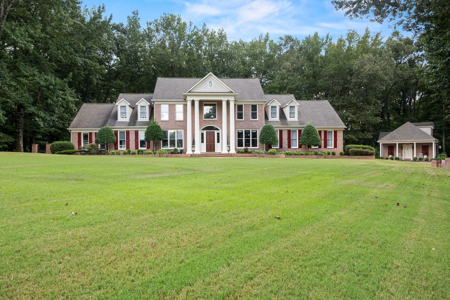 Large white colonial style mansion with pillars sits on expansive green lawn surrounded by trees and landscaping.